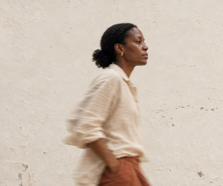 A woman in cream linen and rust trousers walking past a weathered plaster wall